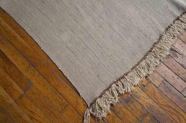 A coarse, flatwoven, light beige rug with subtle stripes and thick fringe rests diagonally on distressed wooden floors.
