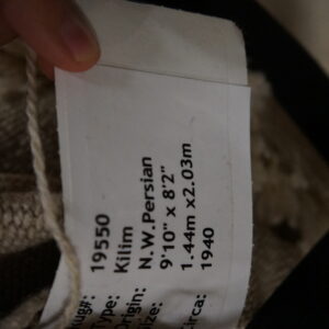 Close-up of a rug tag placed against a tightly woven beige textile showing visible cream fringe.