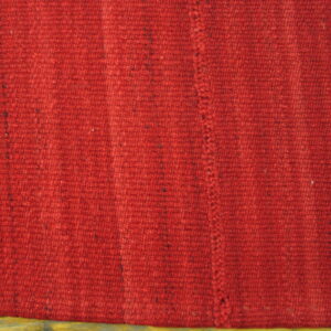 Textured red rug with dark flecks shows vertical ribbing, a stitched seam, and wood flooring below.