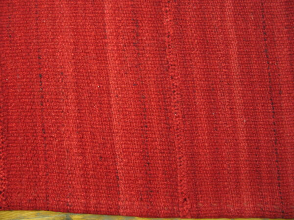 Textured red rug with dark flecks shows vertical ribbing, a stitched seam, and wood flooring below.