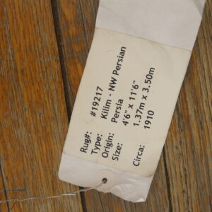 A cream-colored rug information tag, secured by wire, rests on heavily grained wooden flooring.