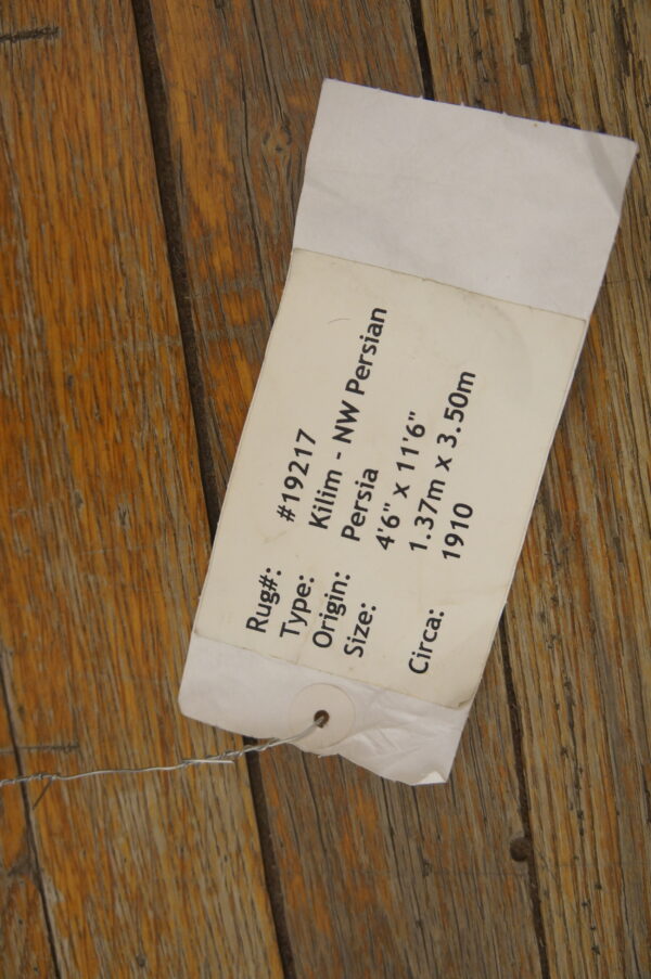 A cream-colored rug information tag, secured by wire, rests on heavily grained wooden flooring.