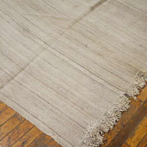 A coarse, light tan flatweave rug corner with shaggy fringe lies on distressed brown wood flooring.