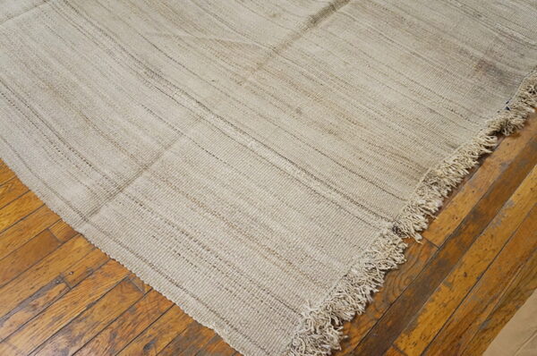 A coarse, light tan flatweave rug corner with shaggy fringe lies on distressed brown wood flooring.
