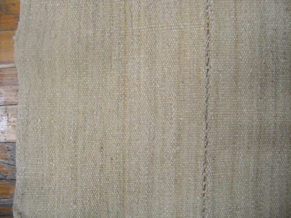 Beige woven rug with a tight texture and stitched vertical line resting on wood planks.