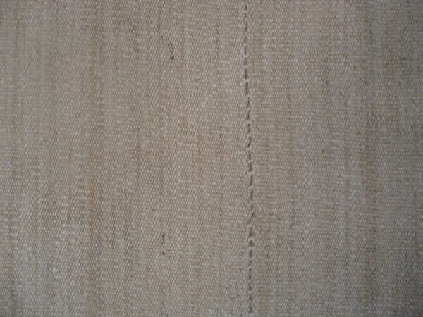 Tightly woven, textured beige rug surface featuring a visible vertical seam of thick, hand-stitched thread.