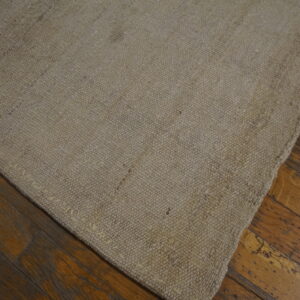 A corner of a solid natural beige textured rug sits on dark, worn wooden floorboards.