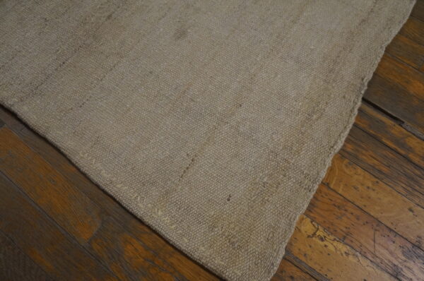 A corner of a solid natural beige textured rug sits on dark, worn wooden floorboards.