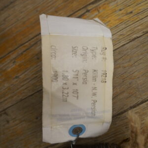 A paper identification tag is attached to the edge of a multicolored rug fragment resting on rustic wooden floorboards.