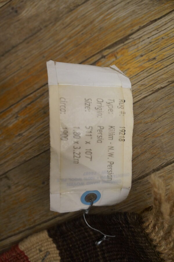 A paper identification tag is attached to the edge of a multicolored rug fragment resting on rustic wooden floorboards.