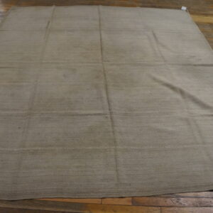 A large, solid beige flatweave rug with horizontal seams rests on rustic brown plank wood flooring.