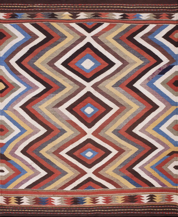 Flatweave rug in red, blue, brown, and cream with bold zigzags forming large central diamonds.