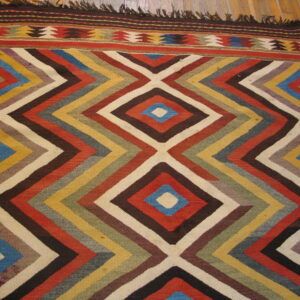Geometric flatweave rug on wood flooring with multicolored zigzags forming large diamond shapes and a striped border.