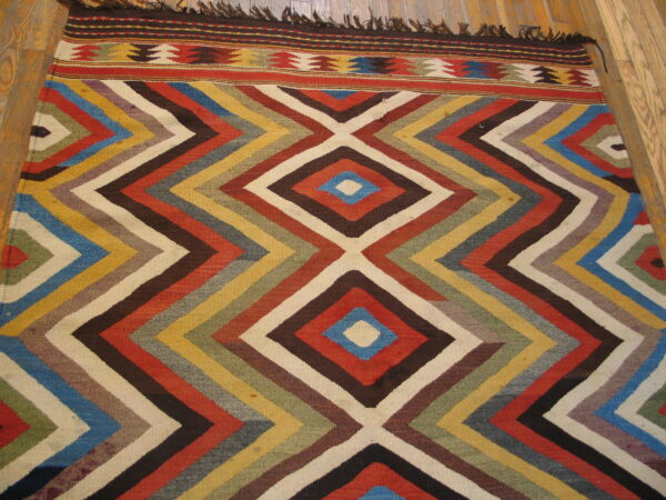 Geometric flatweave rug on wood flooring with multicolored zigzags forming large diamond shapes and a striped border.