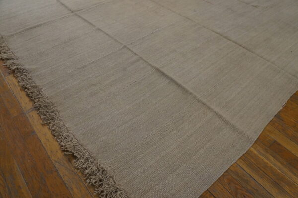 Muted beige flat-woven rug displaying stitched panel construction and fringe on rustic hardwood floors.