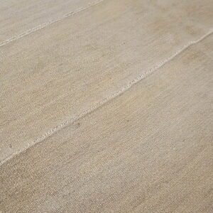 Natural tan rug with a coarse weave texture and strong stitched vertical seams lying on wood.
