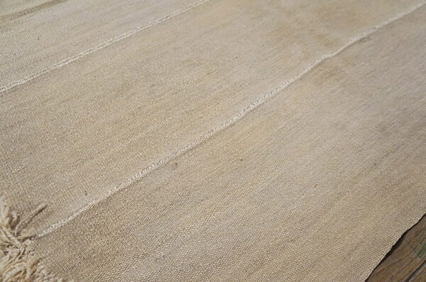 Natural tan rug with a coarse weave texture and strong stitched vertical seams lying on wood.