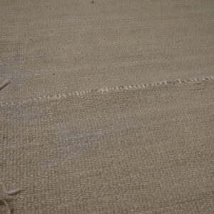 Tan textured rug surface features an irregular stitched seam dividing two woven sections near a shaggy fringe.