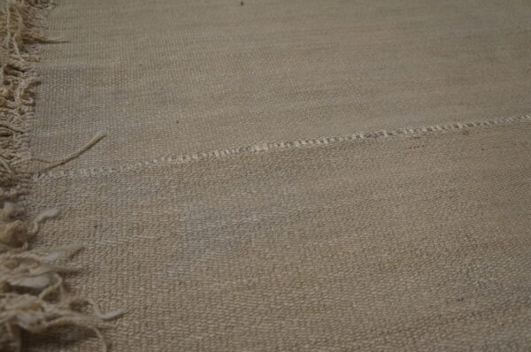 Tan textured rug surface features an irregular stitched seam dividing two woven sections near a shaggy fringe.