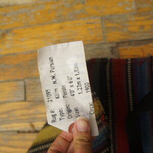 A hand holds a tag over a striped flatweave rug lying on distressed yellow wood floors.