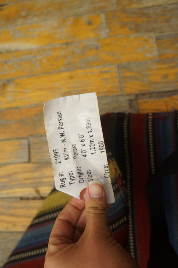 A hand holds a tag over a striped flatweave rug lying on distressed yellow wood floors.