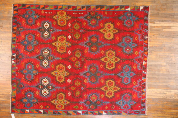 Flat-woven red rug featuring complex blue, mustard, and white geometric medallion patterns on a rustic wood floor.