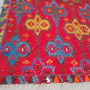 Red rug with geometric blue and gold repeating tribal patterns, visible on a light gray concrete surface.