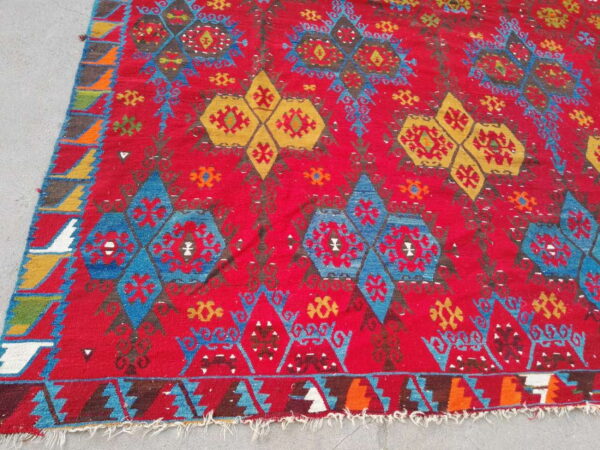 Red rug with geometric blue and gold repeating tribal patterns, visible on a light gray concrete surface.