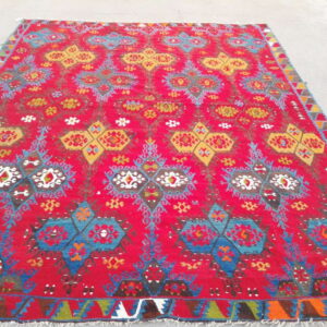 Bright red, low-pile rug featuring complex geometric patterns in yellow, blue, and multicolor accents on a concrete floor.