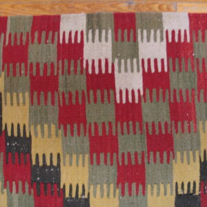Flatweave rug with serrated, interlocking blocks of red, green, yellow, black, and cream on wood flooring.