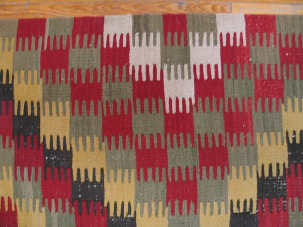 Flatweave rug with serrated, interlocking blocks of red, green, yellow, black, and cream on wood flooring.