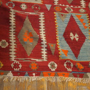 Red flat-weave rug with large tribal diamond motifs in blue, gray, and bright orange colors on wood flooring.