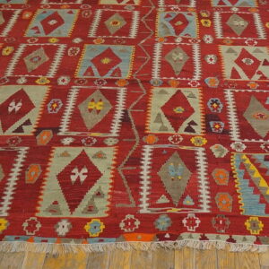 Flatweave rug with red background and geometric diamond shapes in sage green, blue, and gray on wood flooring.