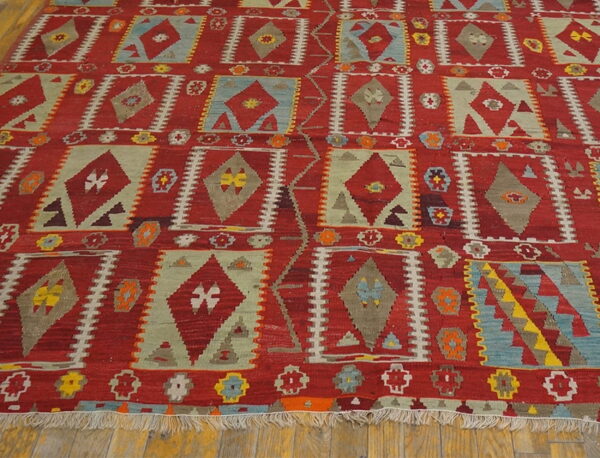 Flatweave rug with red background and geometric diamond shapes in sage green, blue, and gray on wood flooring.