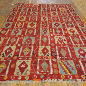 Red rug with geometric diamonds and squares in blue, tan, and khaki lies on a wooden plank floor.