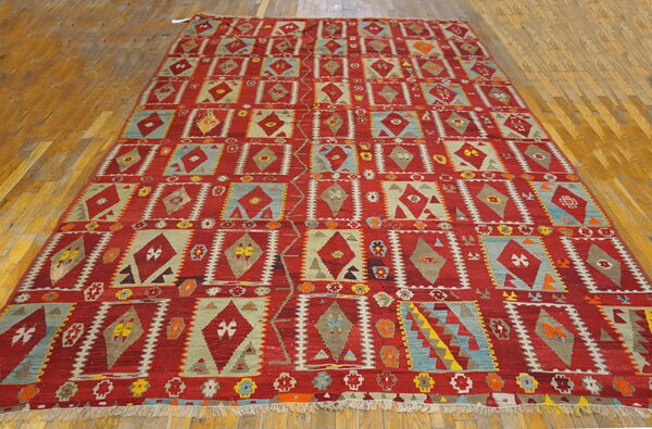 Red rug with geometric diamonds and squares in blue, tan, and khaki lies on a wooden plank floor.