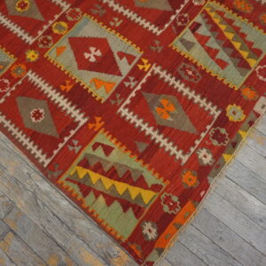Red flatweave rug featuring intricate geometric diamond patterns in sage, gold, and orange on weathered wood.