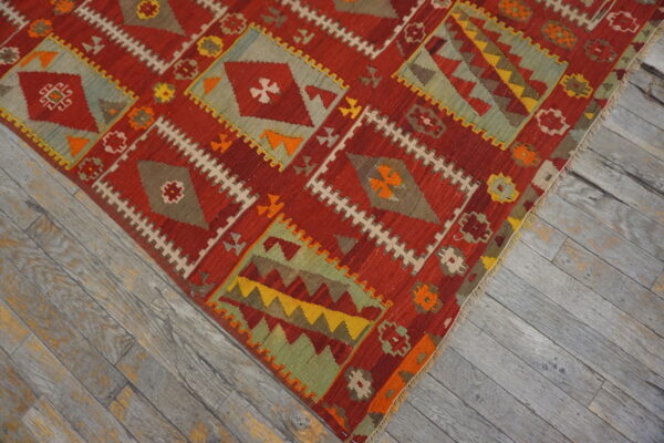 Red flatweave rug featuring intricate geometric diamond patterns in sage, gold, and orange on weathered wood.
