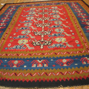 Red, blue, and gold flatweave rug featuring stylized figures and geometric borders on a wood floor.