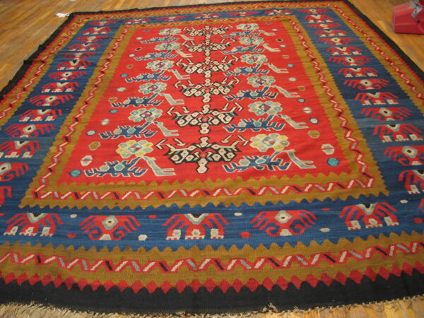 Red, blue, and gold flatweave rug featuring stylized figures and geometric borders on a wood floor.