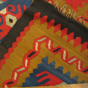 A folded flatweave rug shows serrated, geometric designs in red, blue, black, and mustard gold.
