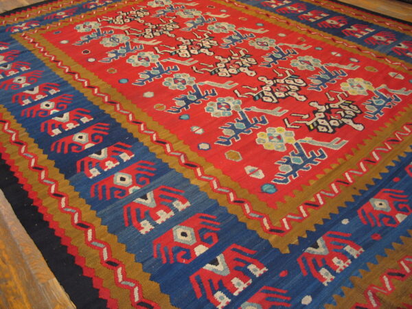 Flat-weave rug with a red center field and blue border featuring stylized figures on wood planks.