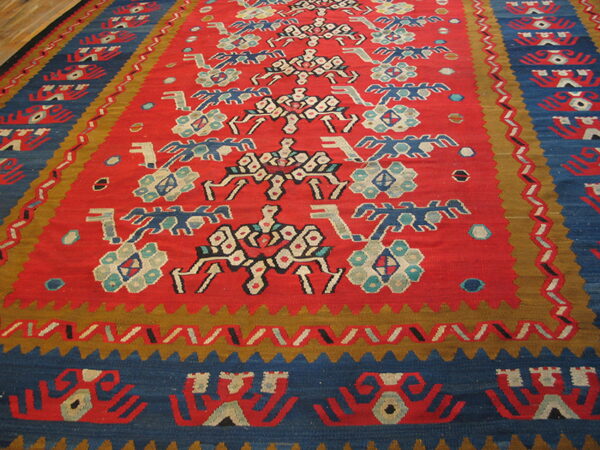 Geometric flatweave rug in red, deep blue, and gold with stylized white and teal tribal motifs.