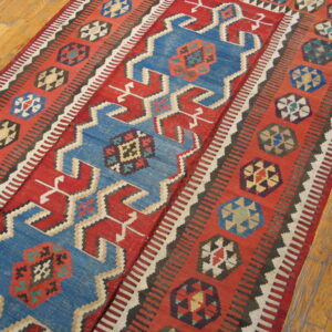 Geometric red kilim rug featuring blue central motifs and multicolored borders on light hardwood flooring.