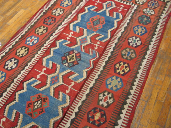 Geometric red kilim rug featuring blue central motifs and multicolored borders on light hardwood flooring.