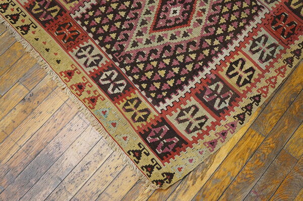 Flatweave rug with pink, red, brown, and khaki geometric patterns rests on worn wooden floorboards.