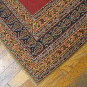 Flat-woven rug corner shows a red field surrounded by detailed geometric borders in navy, yellow, and red on wood flooring.