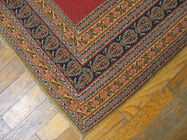 Flat-woven rug corner shows a red field surrounded by detailed geometric borders in navy, yellow, and red on wood flooring.