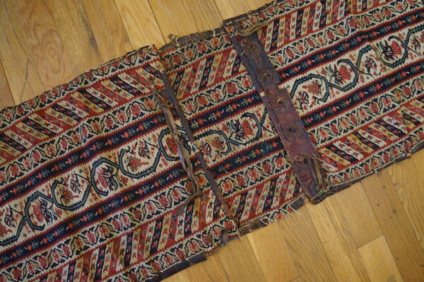 Segmented red, navy, and cream flatweave textile with stylized patterns and weathered leather straps on hardwood.