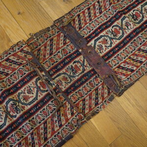 Geometric red, blue, and cream banded textile piece featuring dark leather straps on wooden floor.